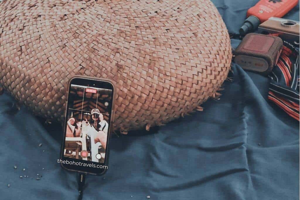 Smartphone recording a video while leaning against a woven straw hat on dark fabric with camera accessories nearby. A behind the scenes view of travel content being filmed with compact travel blogging gear.