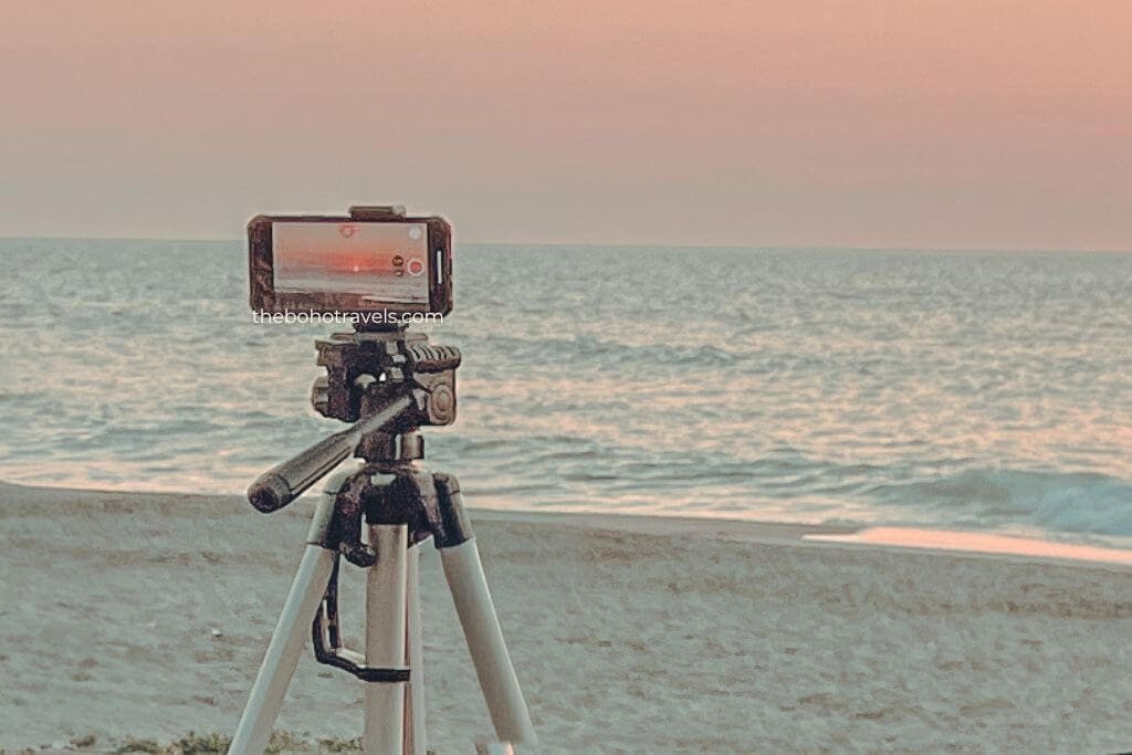 Smartphone mounted on a tripod recording the sunset over a calm beach with gentle waves in the background. A portable setup for filming travel videos using lightweight travel blogging gear.