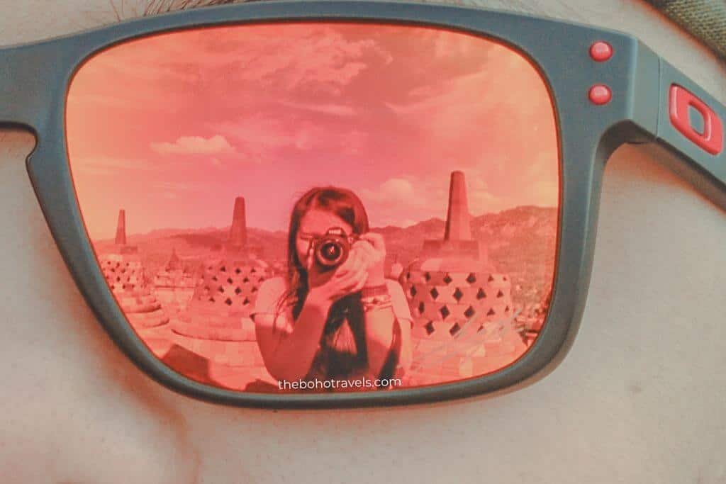 Reflection of a person holding a camera captured in the red mirrored lens of a pair of sunglasses with temple structures visible in the background. A creative travel photography perspective using everyday accessories.