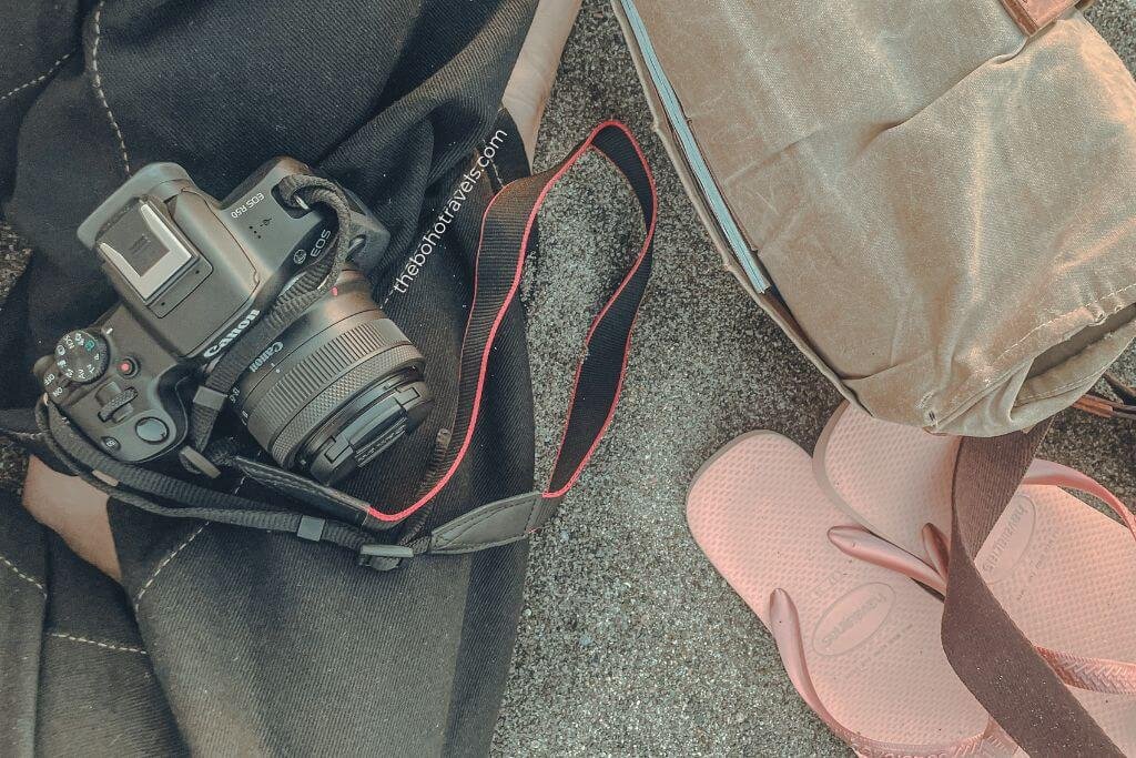 Canon camera resting on a black bag beside a backpack and pink flip flops on the ground. A flat lay of essential travel blogging gear packed for a day of exploring and content creation.